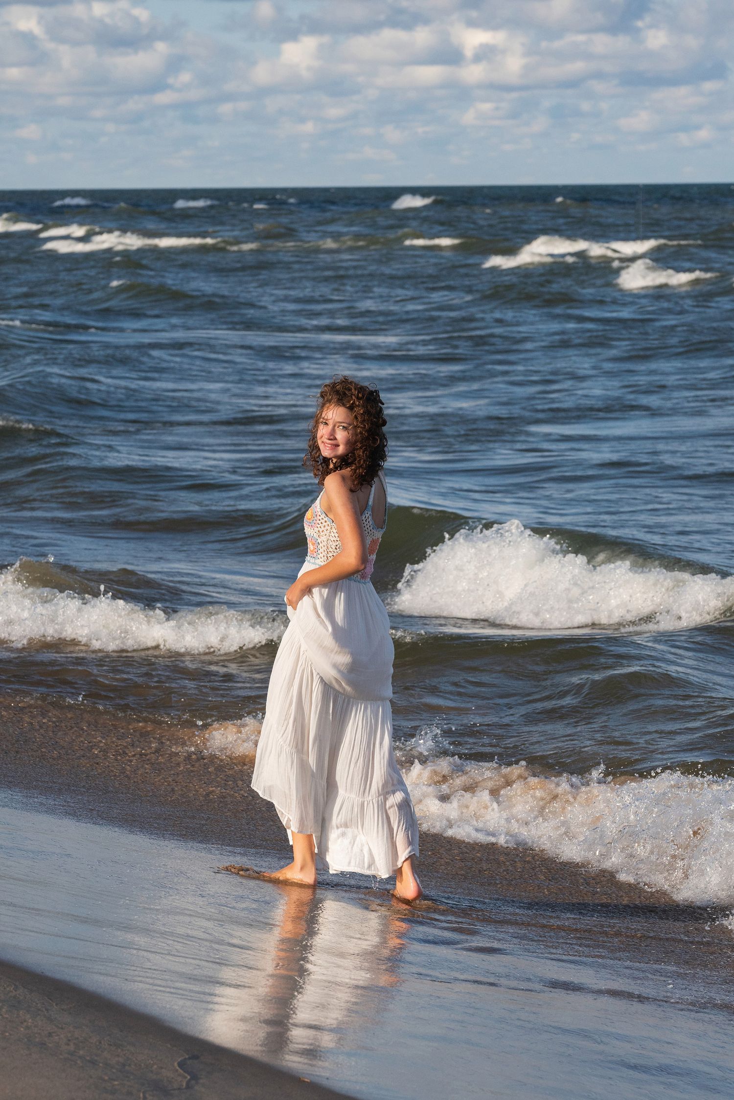 Person in flowing white dress dances with ocean waves on a sunny beach.