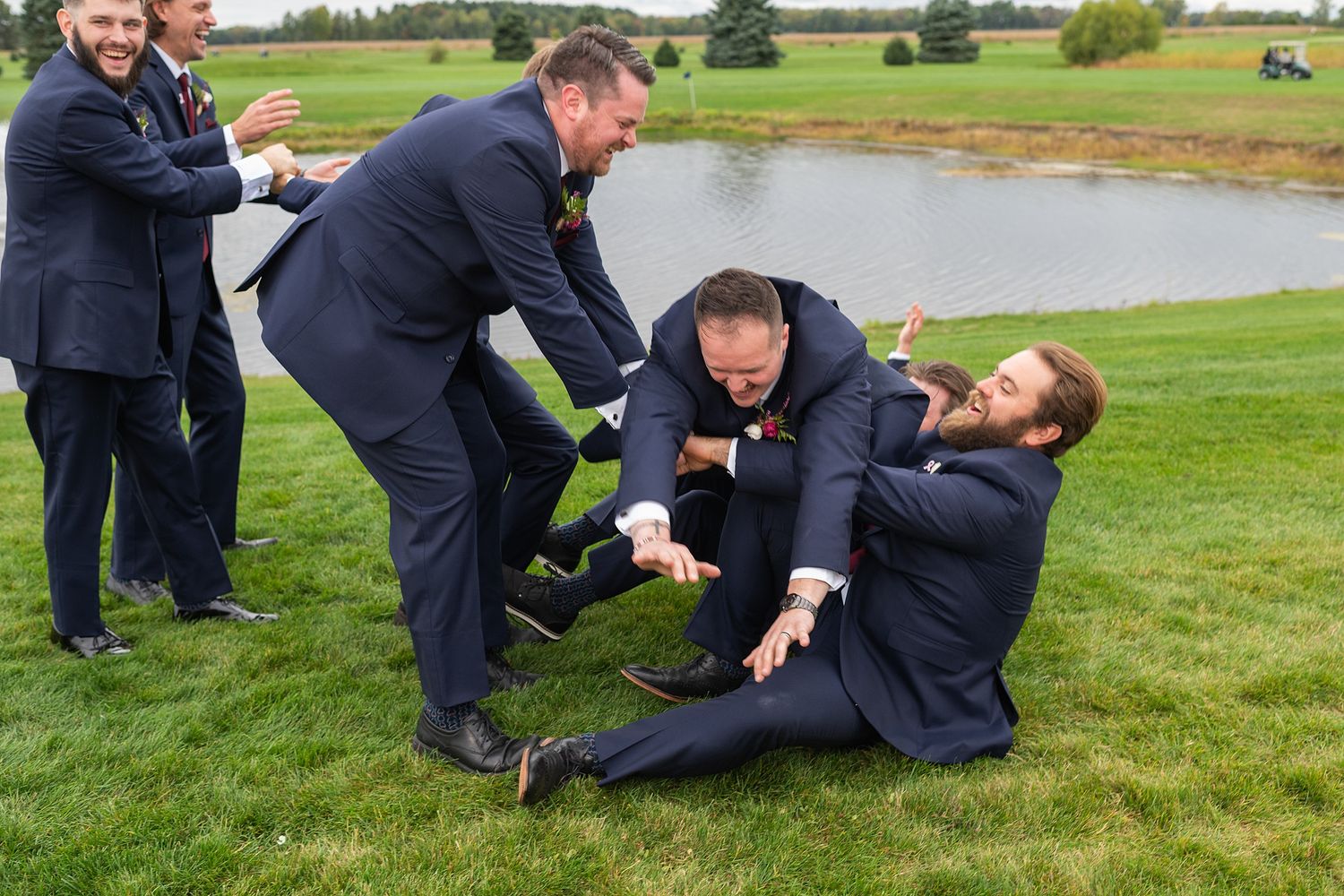 Groomsmen in navy suits playfully wrestling on green grass near a pond at a wedding venue.