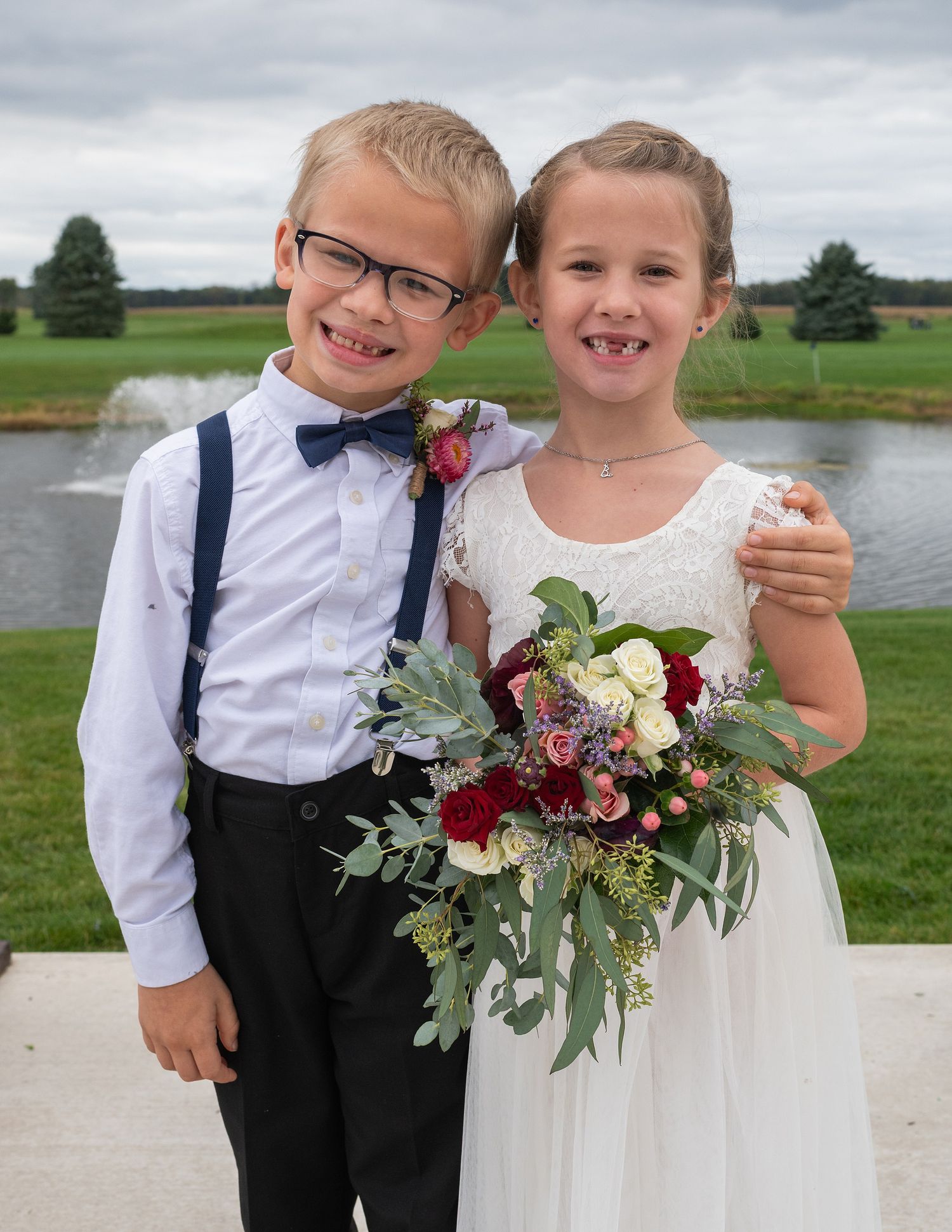 Two young children dressed in wedding attire, boy in suspenders and bow tie, girl in white dress holding bouquet.