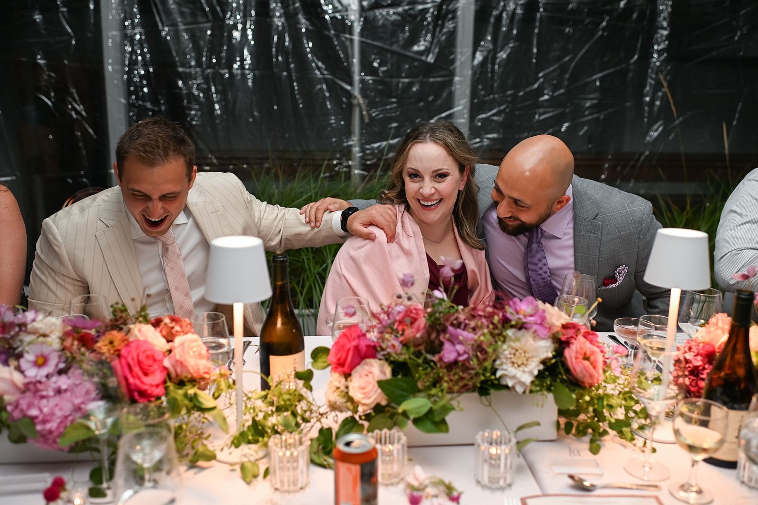 Wedding guests gather around an elegantly decorated dinner table with pink floral centerpieces and glowing candlelight.