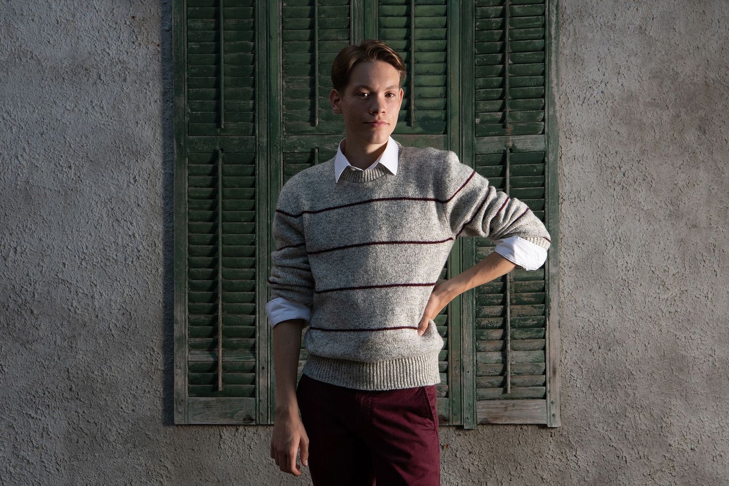 Person in striped sweater poses against weathered wall with green shutters.