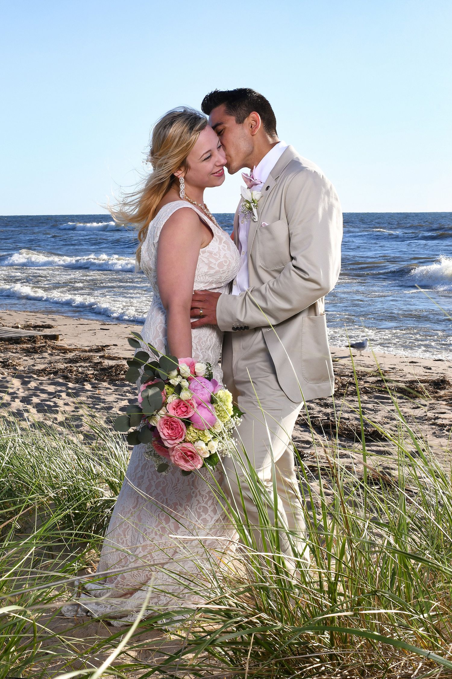 Romantic beach wedding portrait with pink bouquet against ocean backdrop.