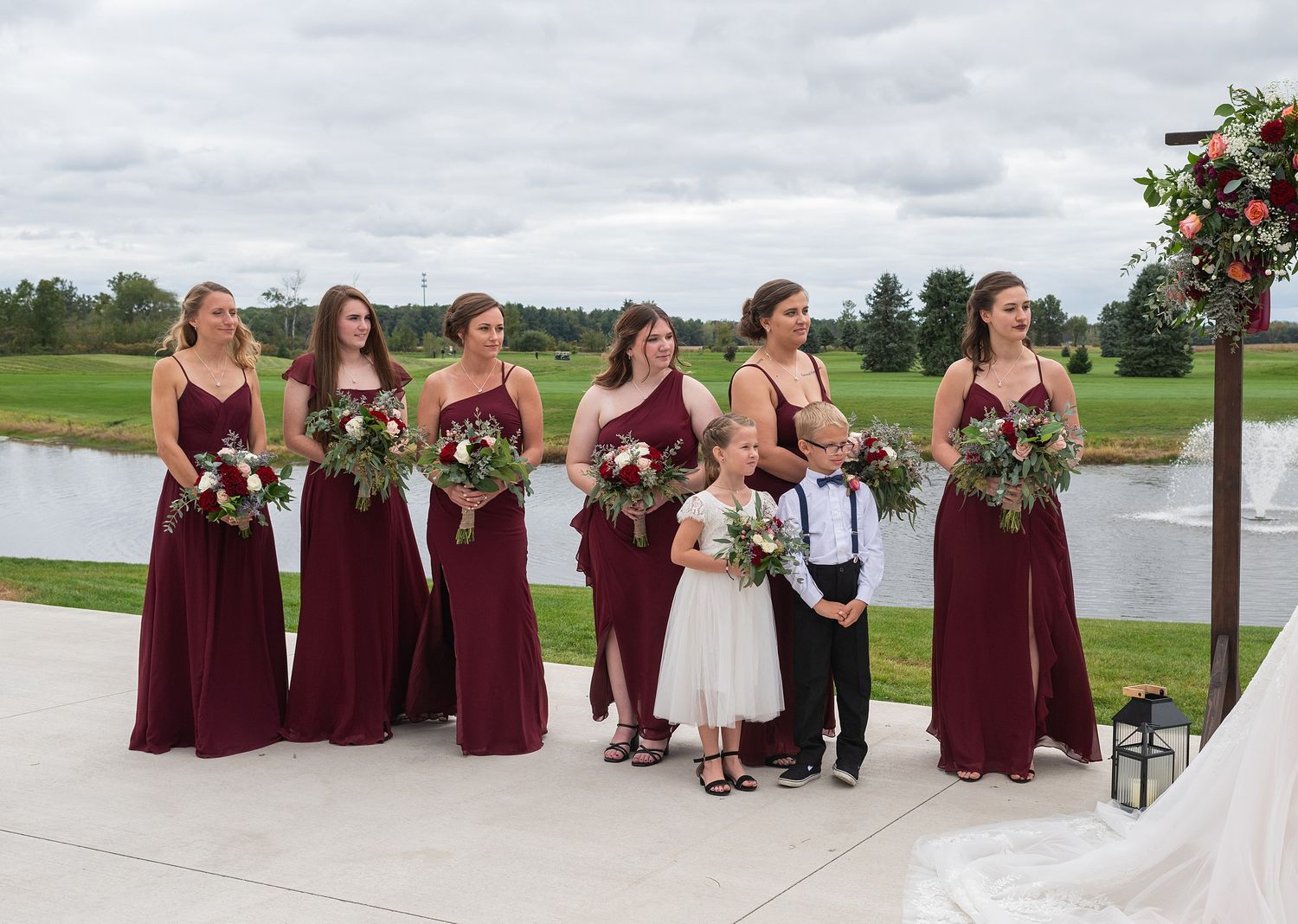 Six bridesmaids in burgundy gowns holding floral bouquets stand outdoors at a wedding ceremony near a scenic pond.