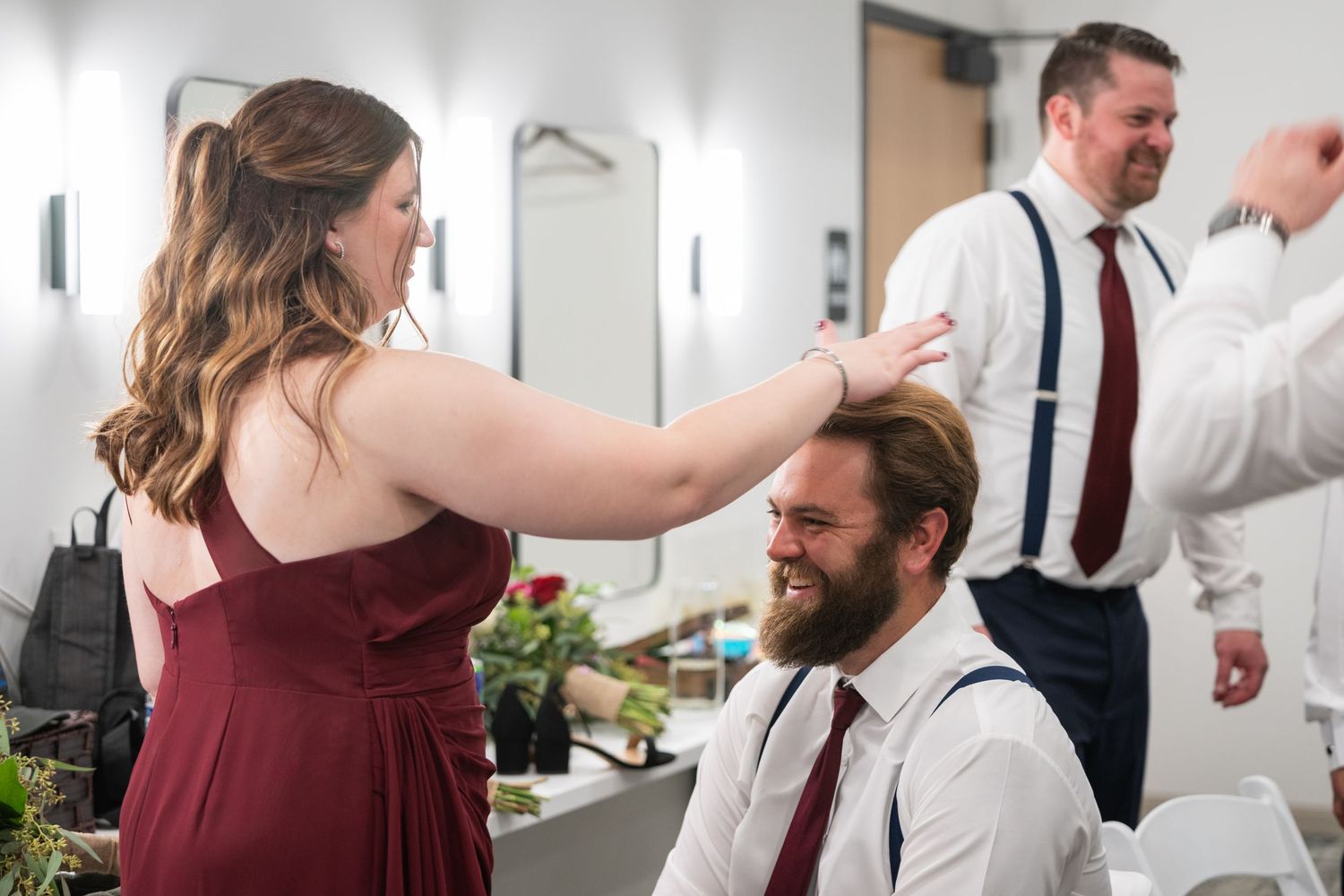 Wedding party getting ready, woman styling groomsman's hair while man in suspenders adjusts tie in mirror.