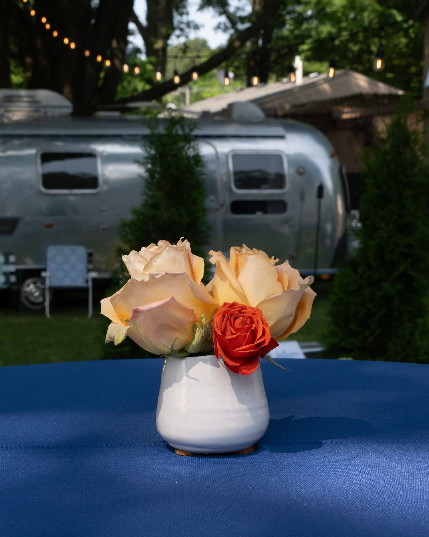 Peach and coral roses in a white vase sit on a blue tablecloth with a silver Airstream trailer visible in the background.