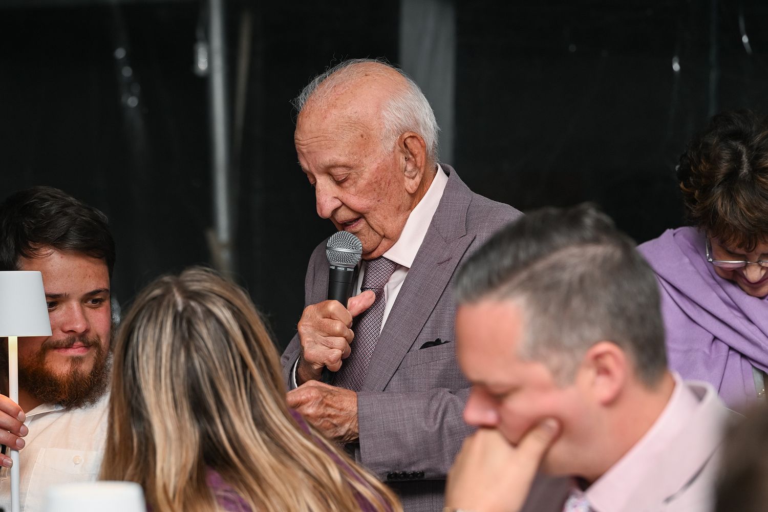 A person in a gray suit speaks into a microphone while others sit at a dining table during what appears to be an event.