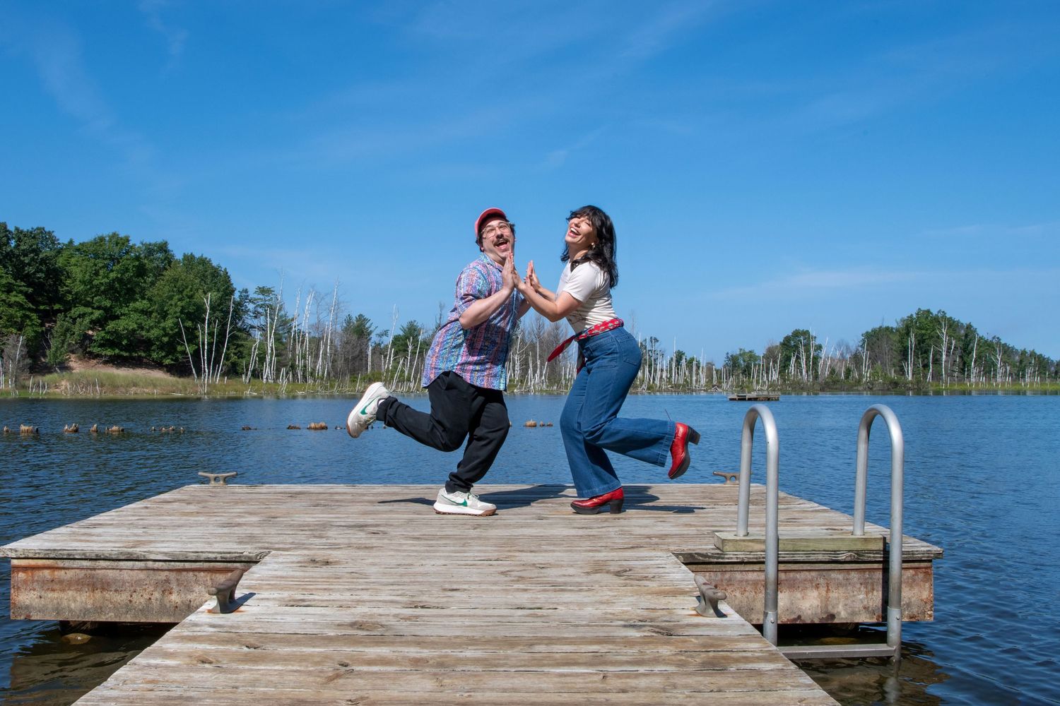 Two people dancing joyfully on a wooden dock by a calm lake under a bright blue sky.