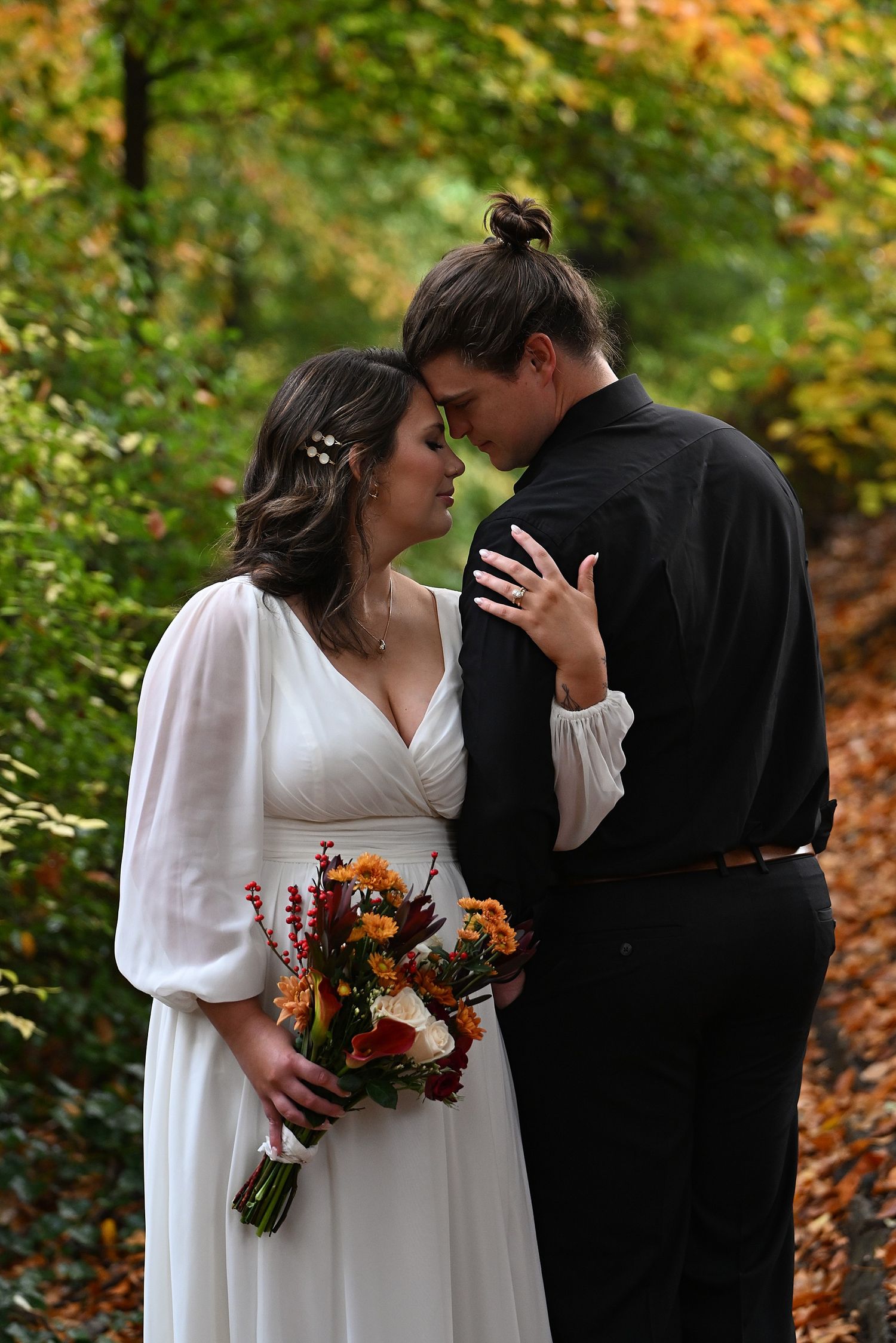 Bride in a white flowy dress holding an orange autumn bouquet touches foreheads with her groom in black among colorful fall foliage.