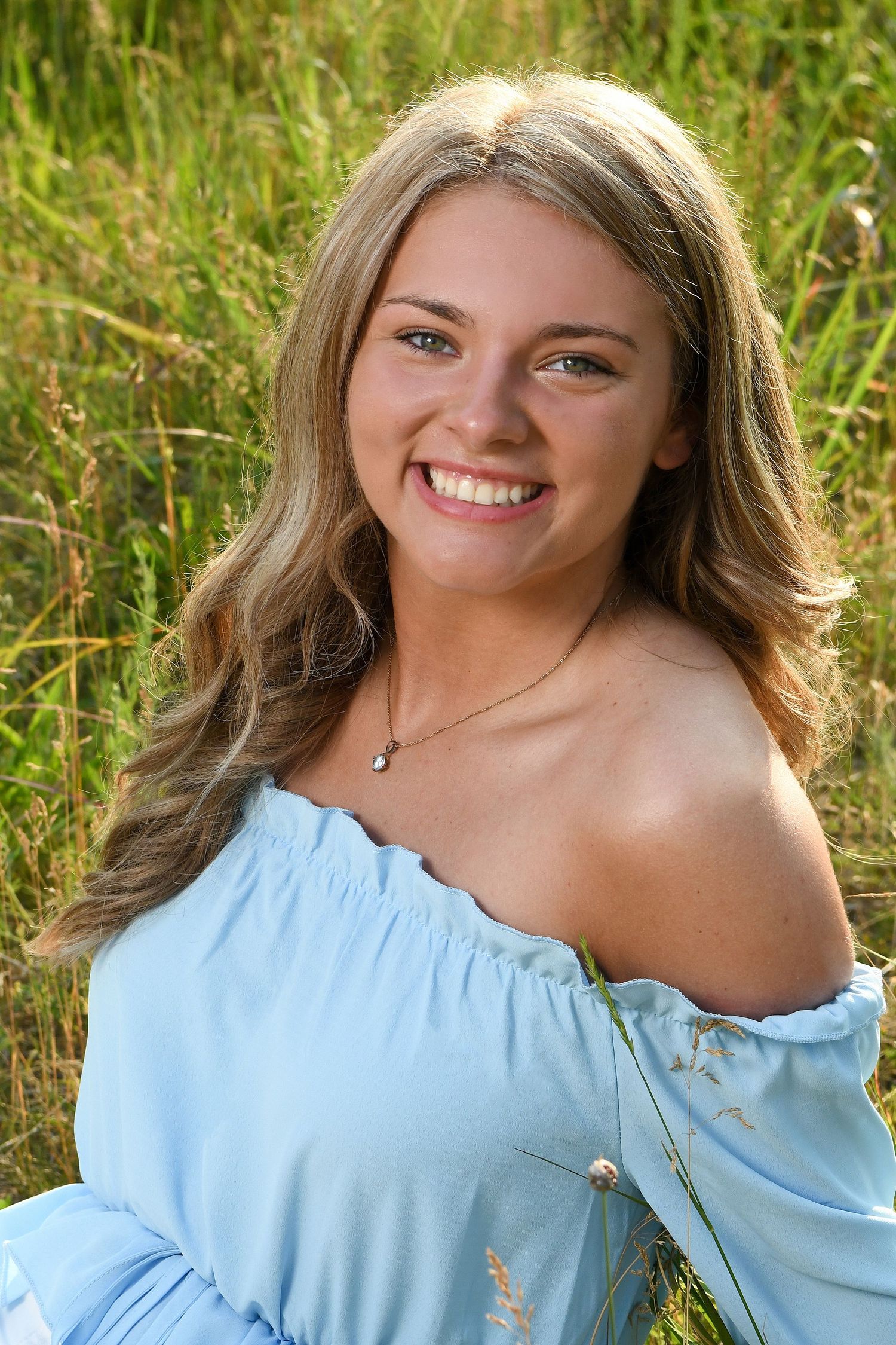 Woman in light blue off-shoulder top poses in field of tall grass.