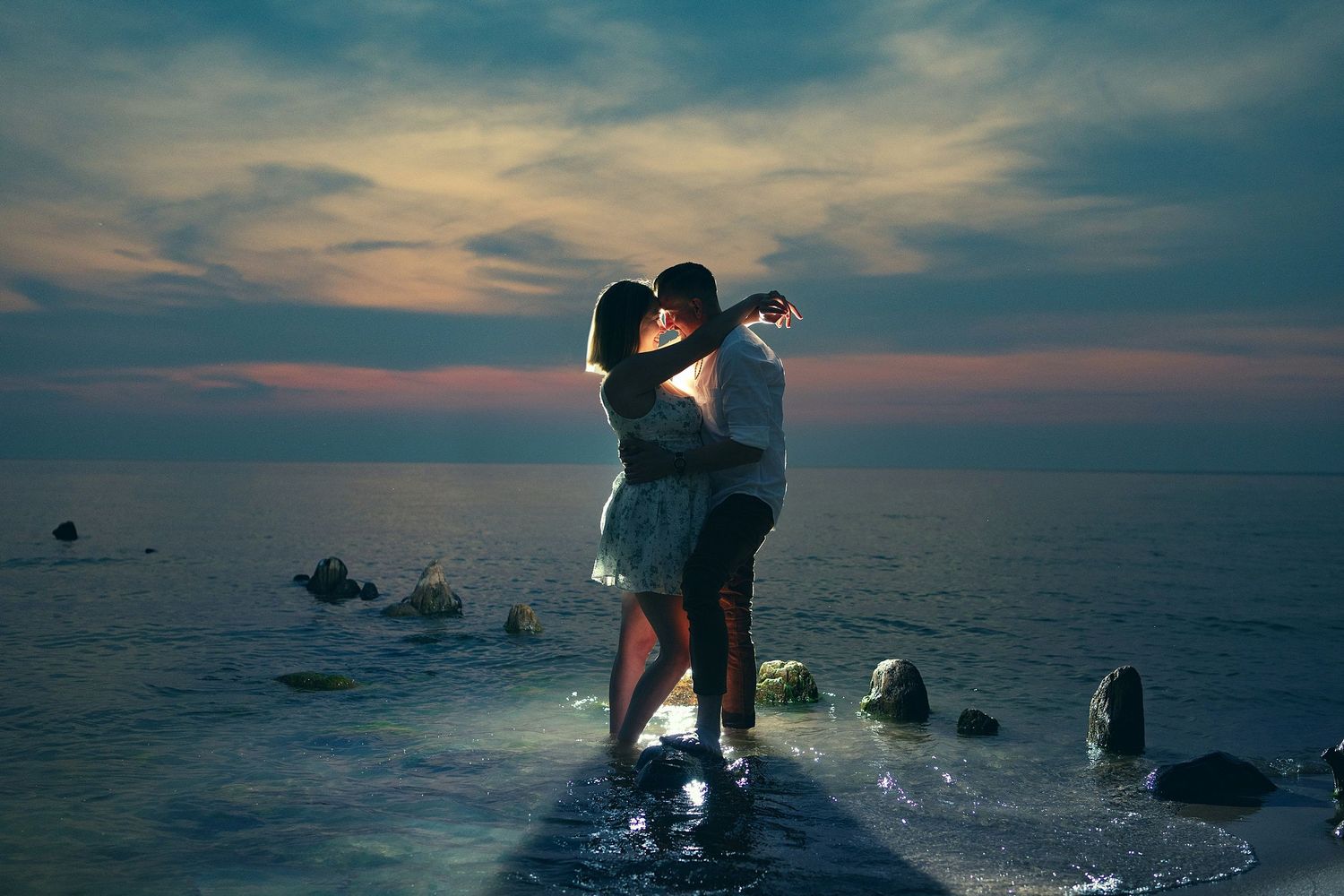 Romantic sunset silhouettes by ocean with rocks in foreground.