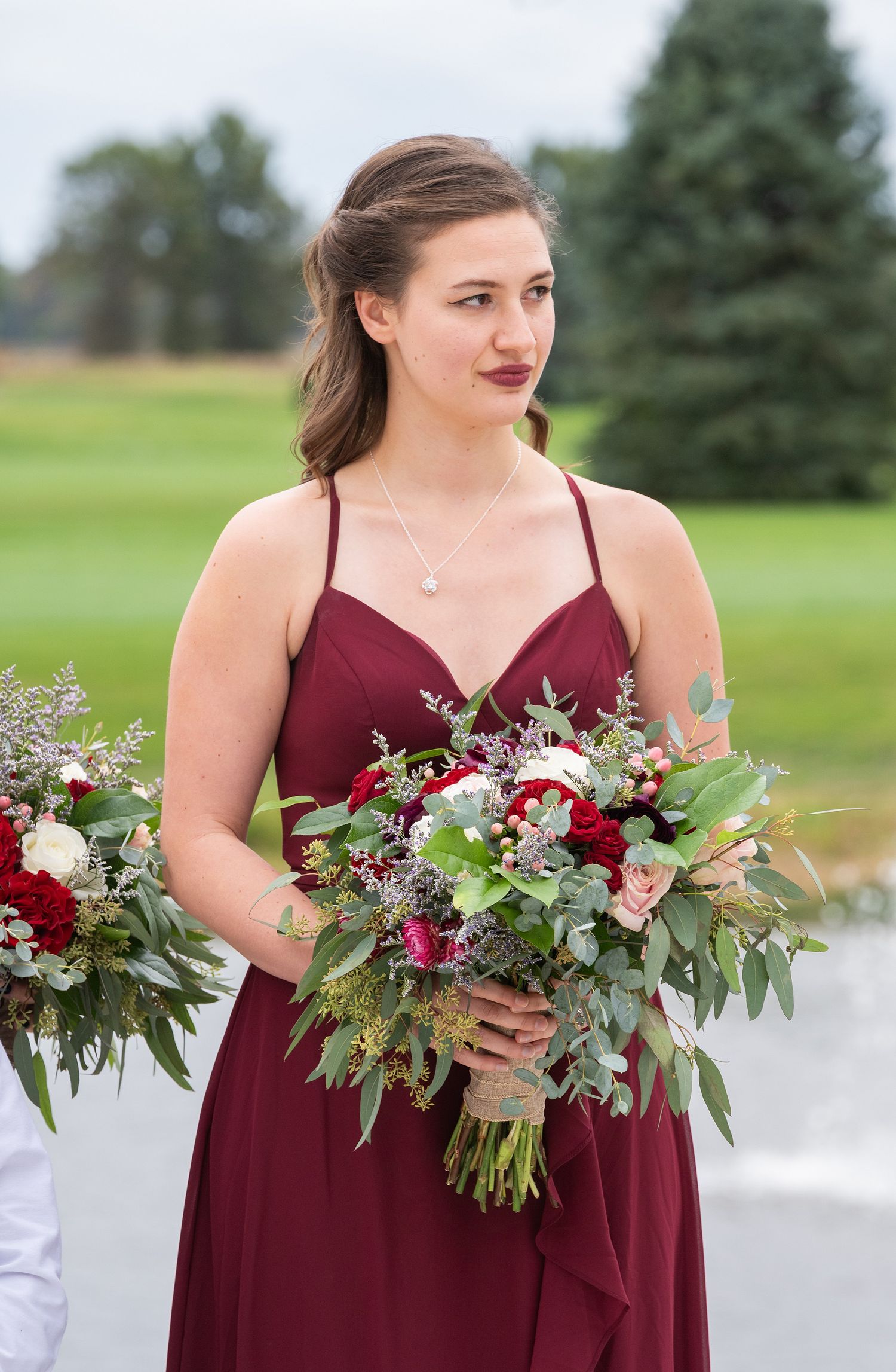 Bridesmaid in burgundy dress holding lush red and green floral bouquet at outdoor wedding ceremony.