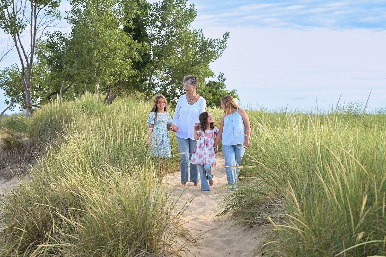Family walking on sandy beach path through tall grass dunes on a sunny summer day.