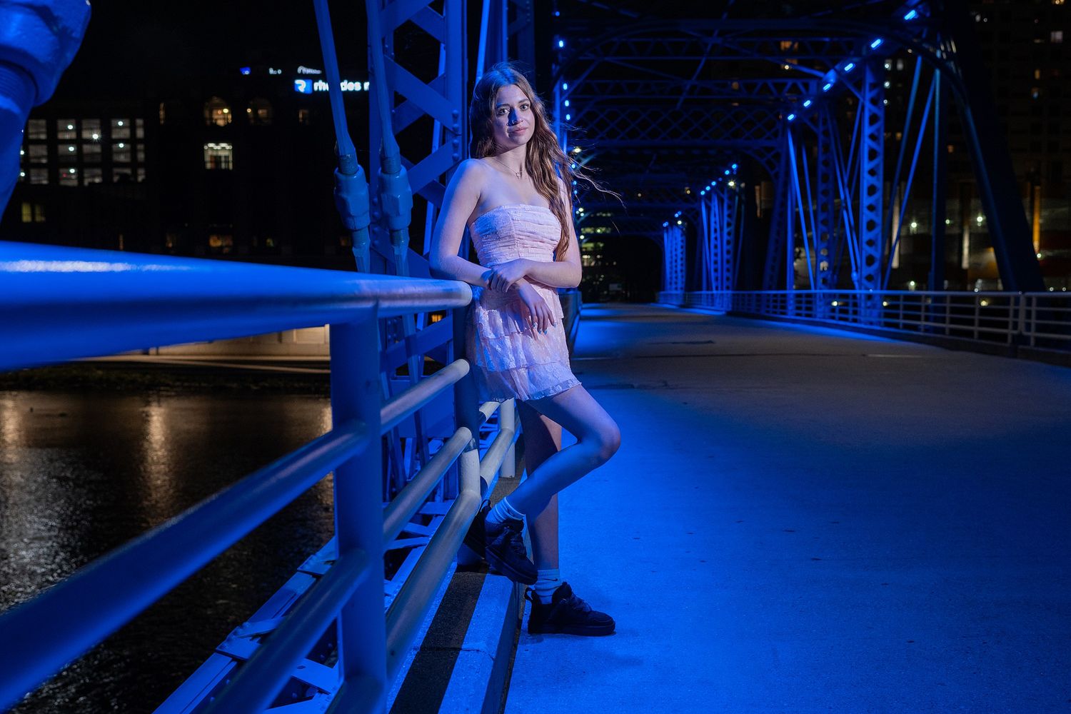 A person leans against a railing on a blue-lit bridge with city lights in the background.