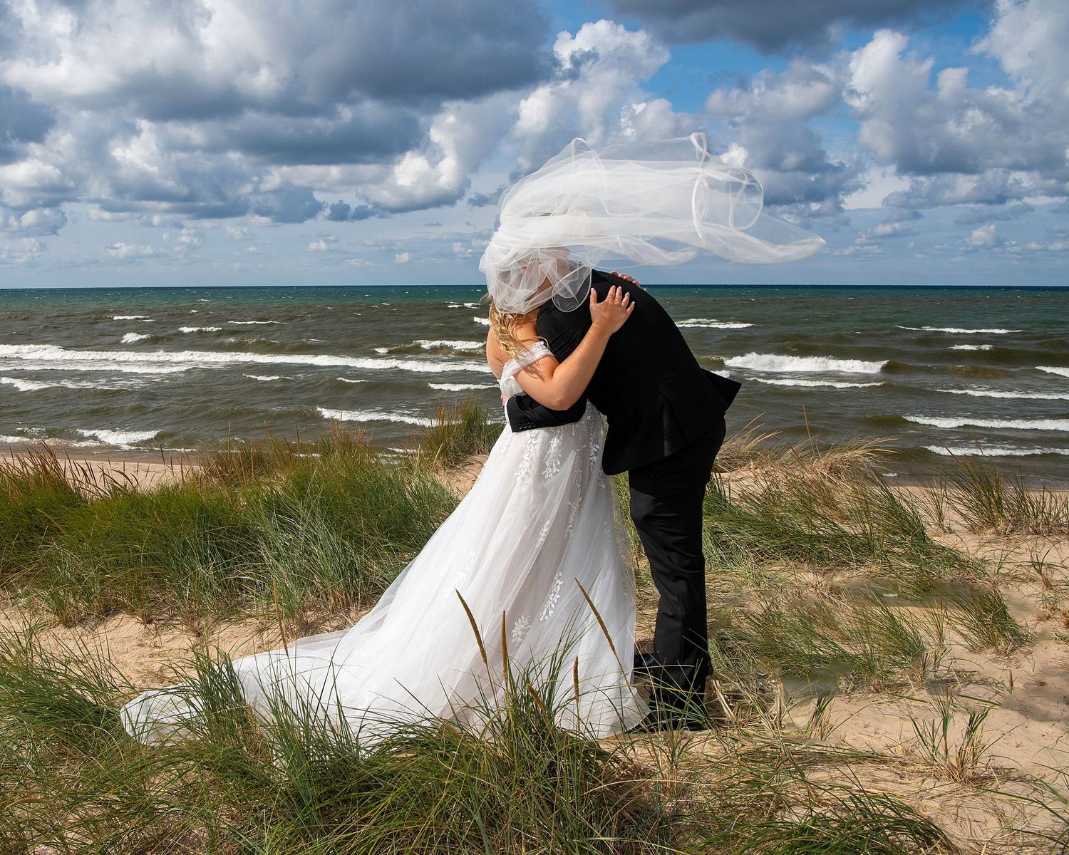 A romantic embrace on a windy beach with waves and clouds creating a dramatic backdrop.