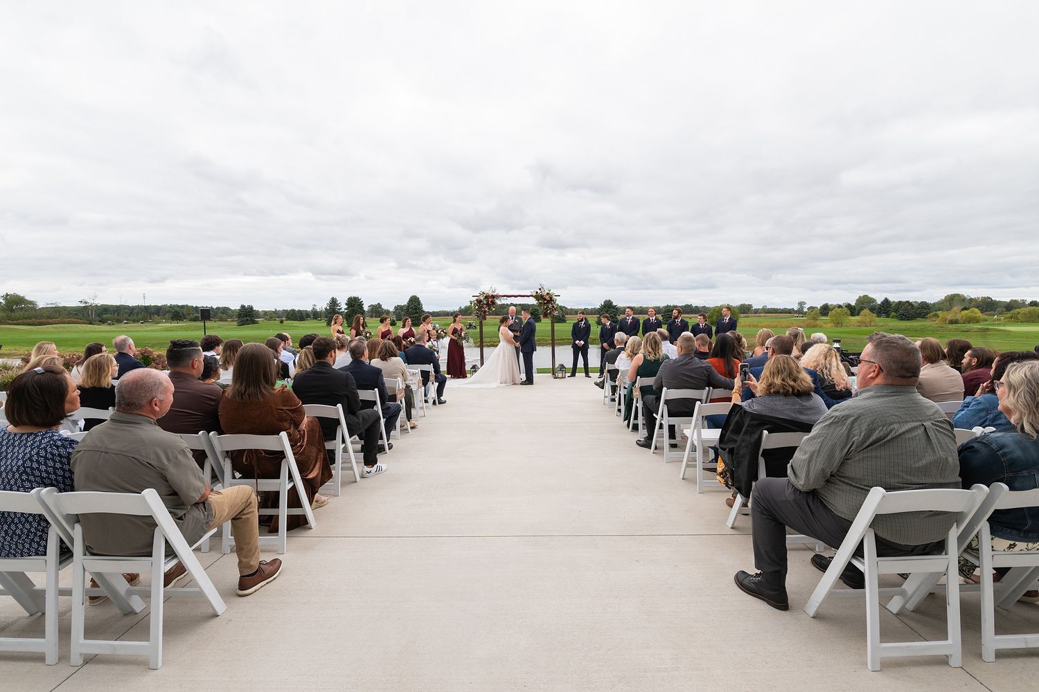 Guests seated in white chairs along an outdoor wedding aisle watch the ceremony under a cloudy sky.
