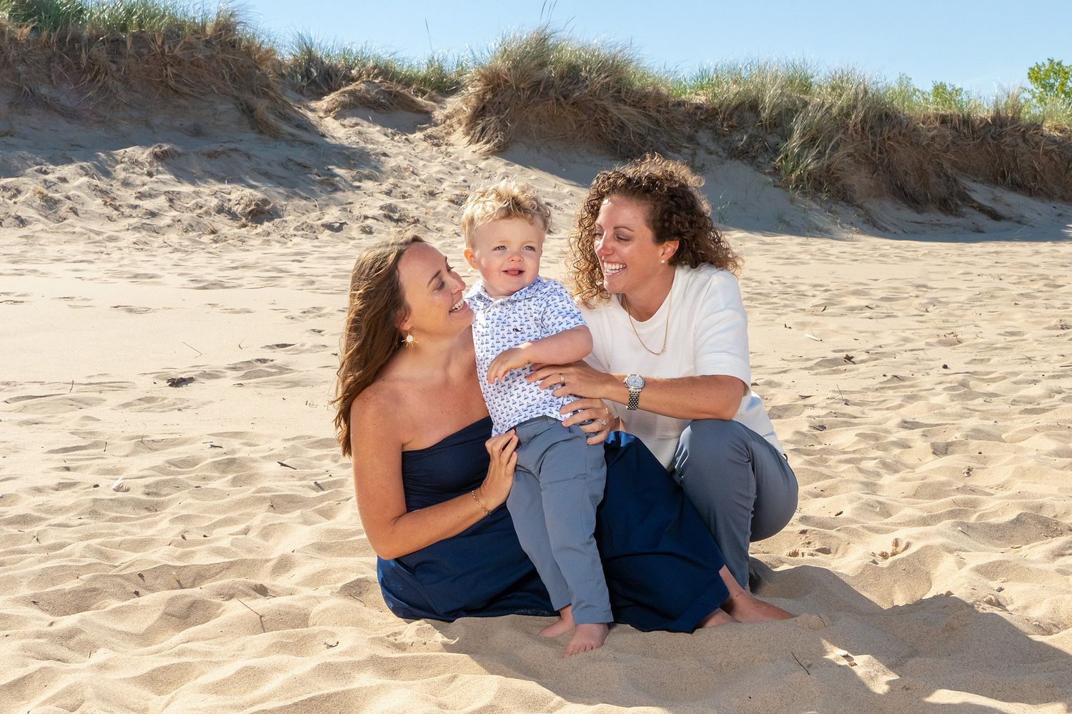 Two women and a baby sit smiling on sandy beach dunes in a warm, sunny family portrait.