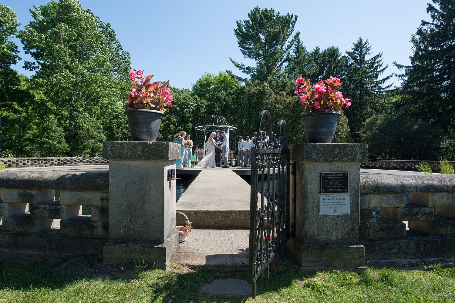 Outdoor wedding ceremony at ornate stone gate entrance with pink floral urns and lush green trees on sunny day.