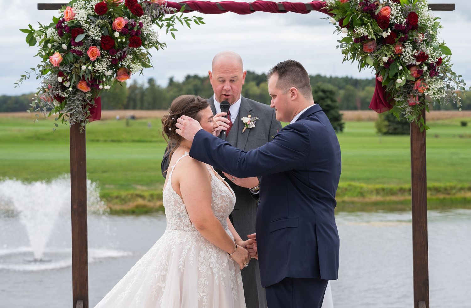 Bride and groom exchange vows under floral arch with officiant at outdoor wedding ceremony near pond.