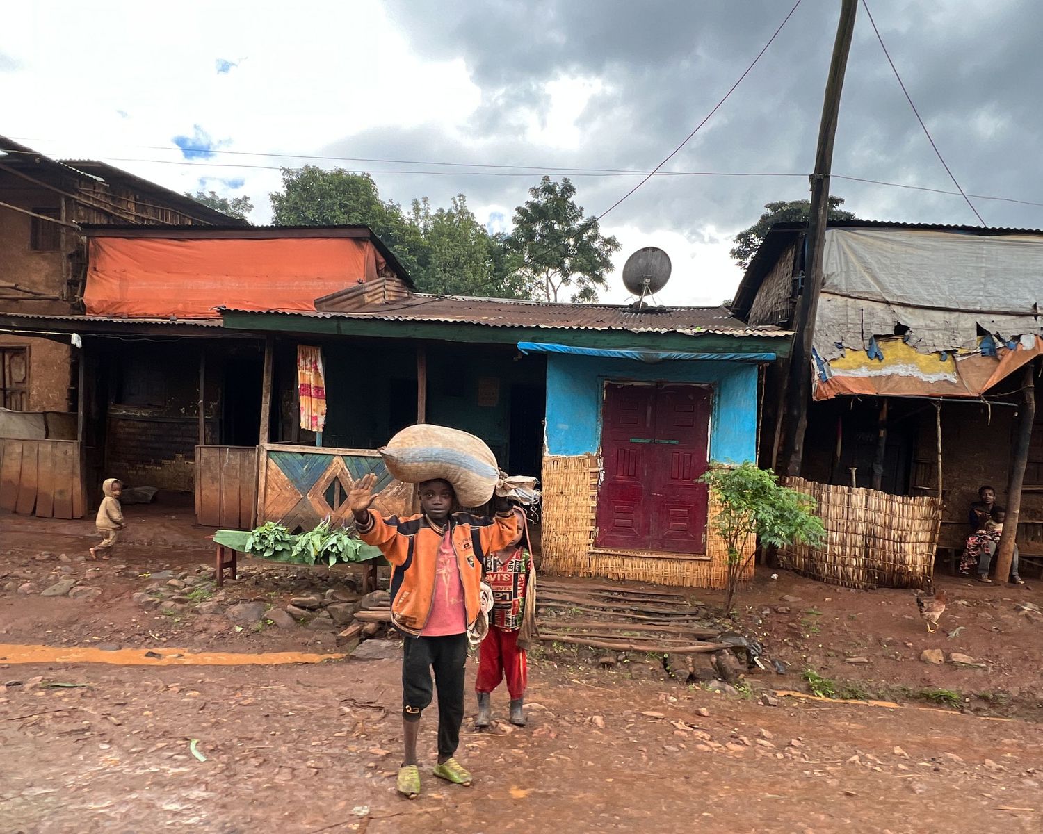 Rural village street with simple buildings and reddish dirt ground under cloudy skies.