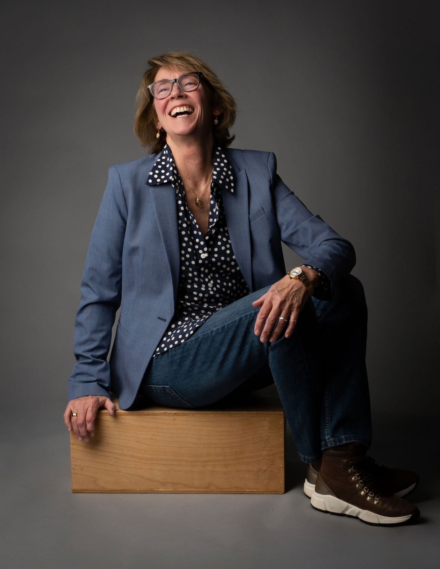 Professional wearing a blue blazer and polka dot blouse sits casually on wooden box while laughing joyfully.