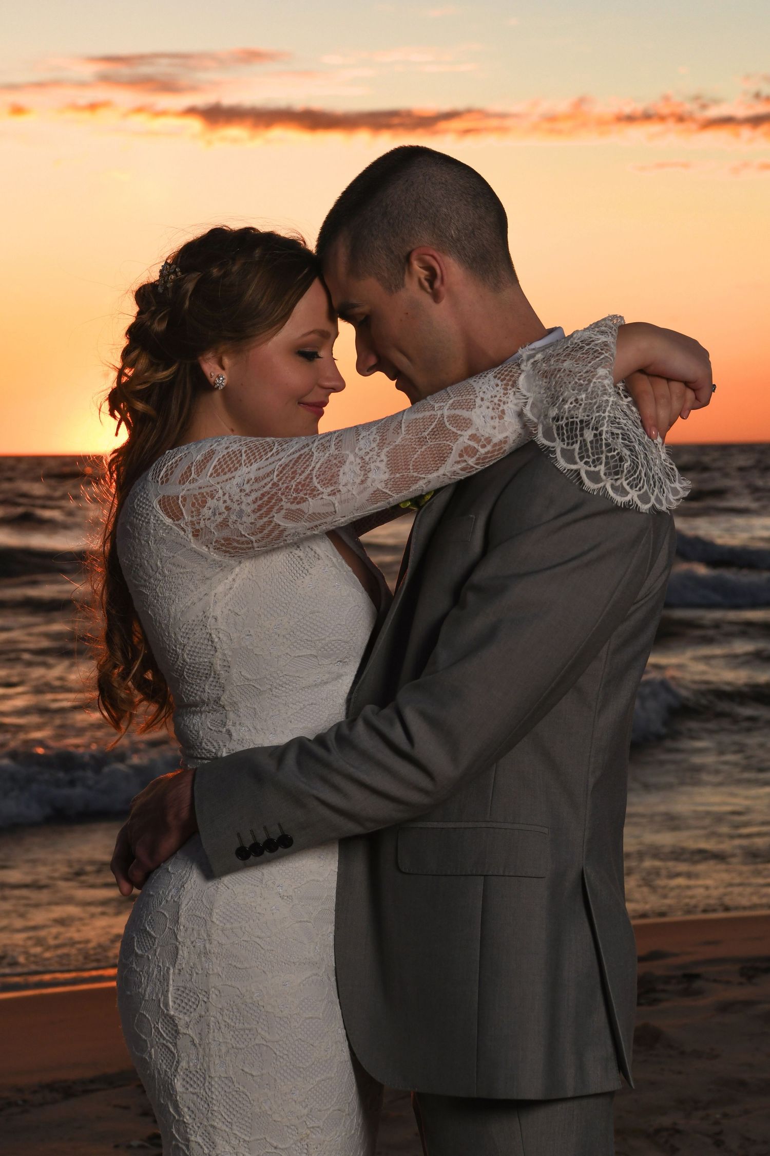 Bride and groom embracing at sunset on the beach, wearing white lace dress and gray suit.