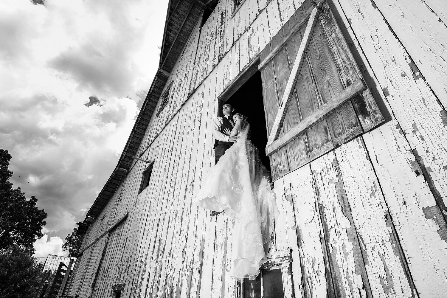 Black and white photo of a bride in a flowing white gown standing in the open doorway of a weathered wooden barn under a cloudy sky.