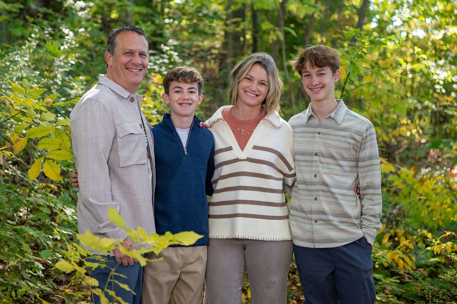 A family of four poses together outdoors against a leafy green background.