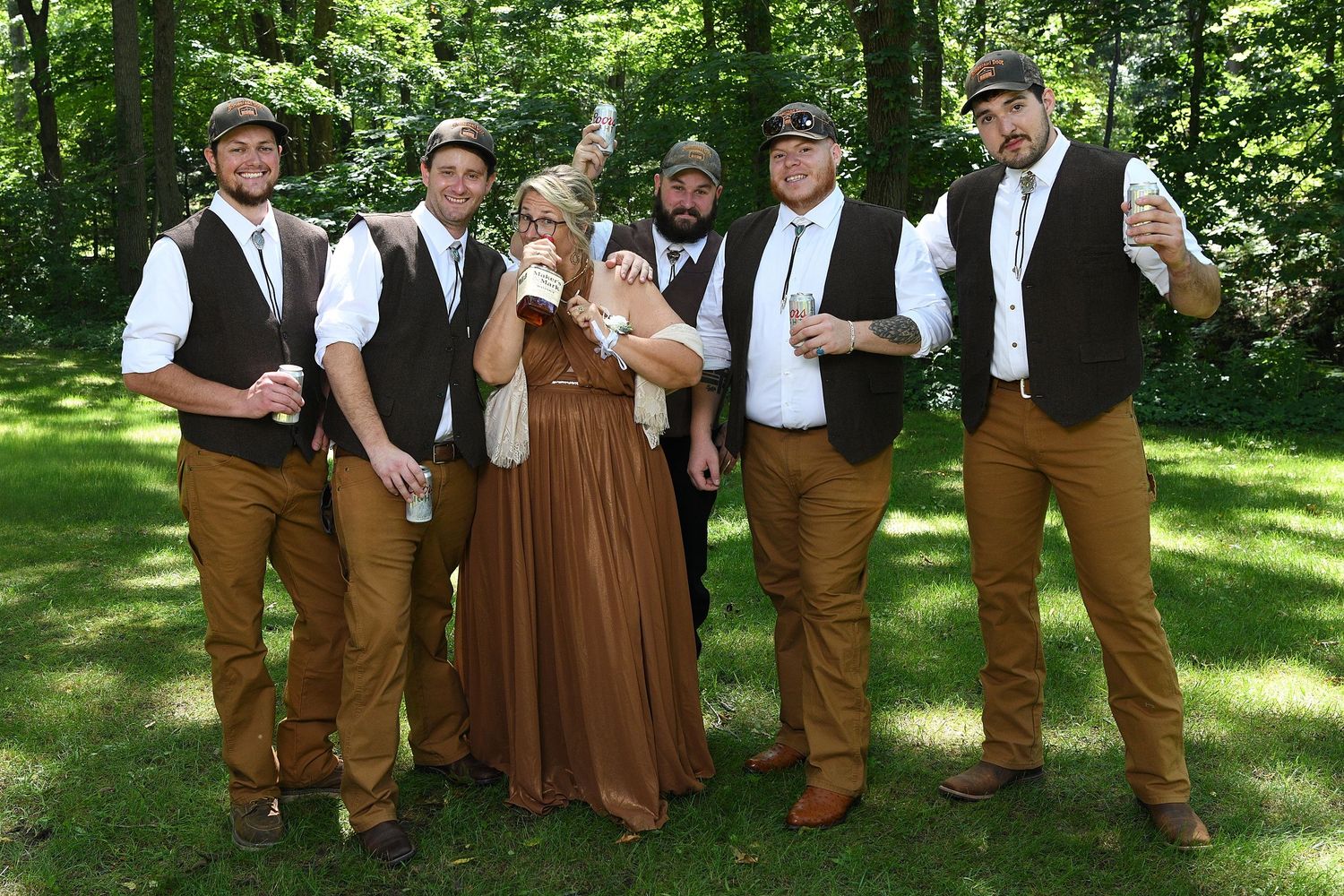 A rustic wedding party in brown vests and suspenders poses together in a sunny garden setting.