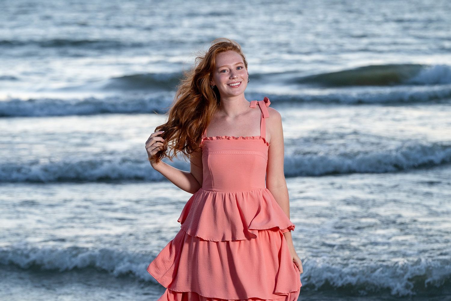 Young woman in pink ruffled dress smiling on beach with ocean waves behind her.