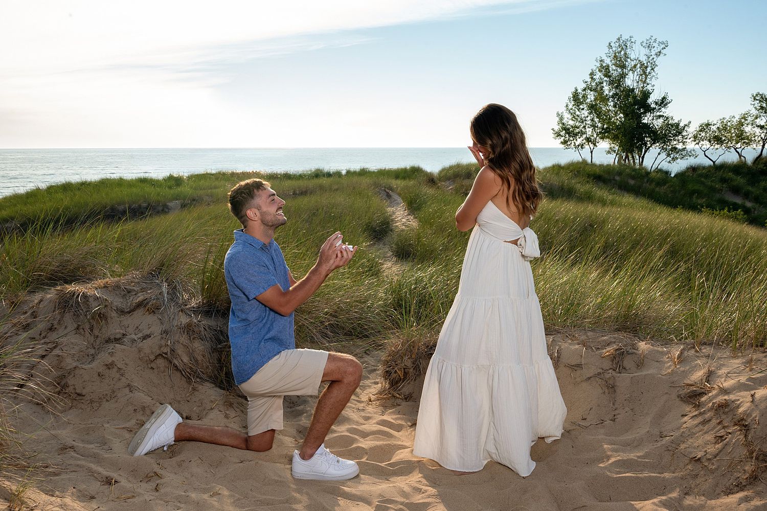 Man proposing to woman in white dress on sandy beach dunes at sunset near ocean.