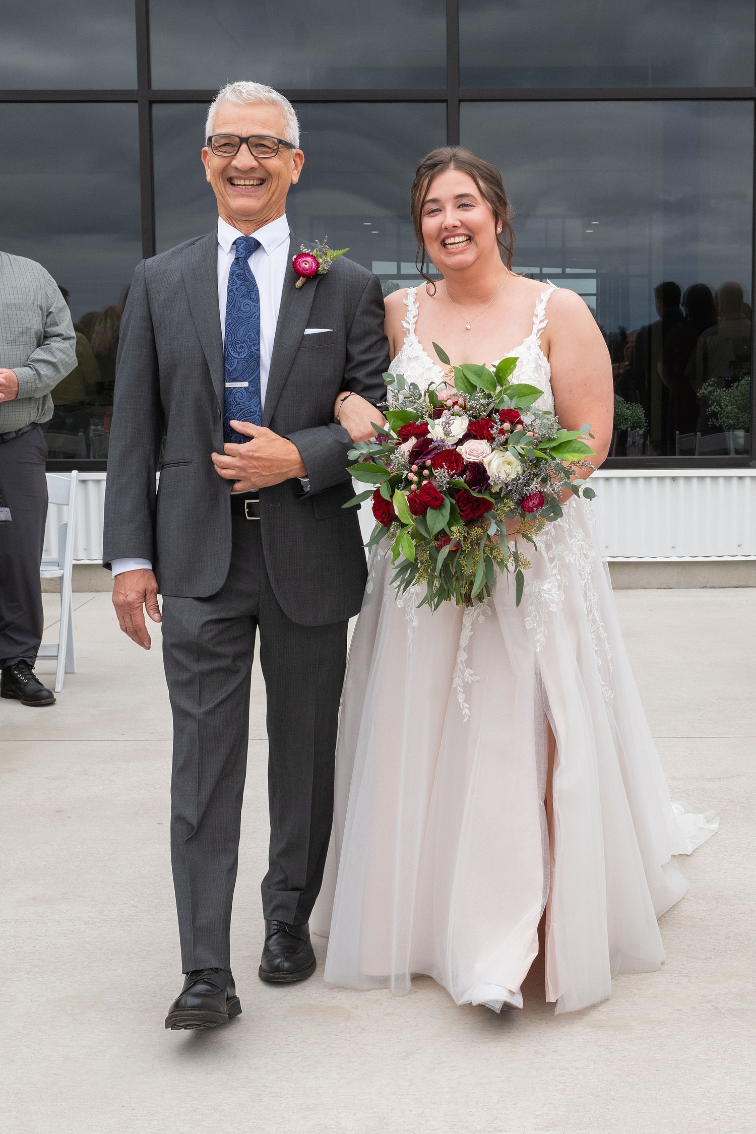 Smiling father walks bride in white lace gown holding red floral bouquet down the aisle.