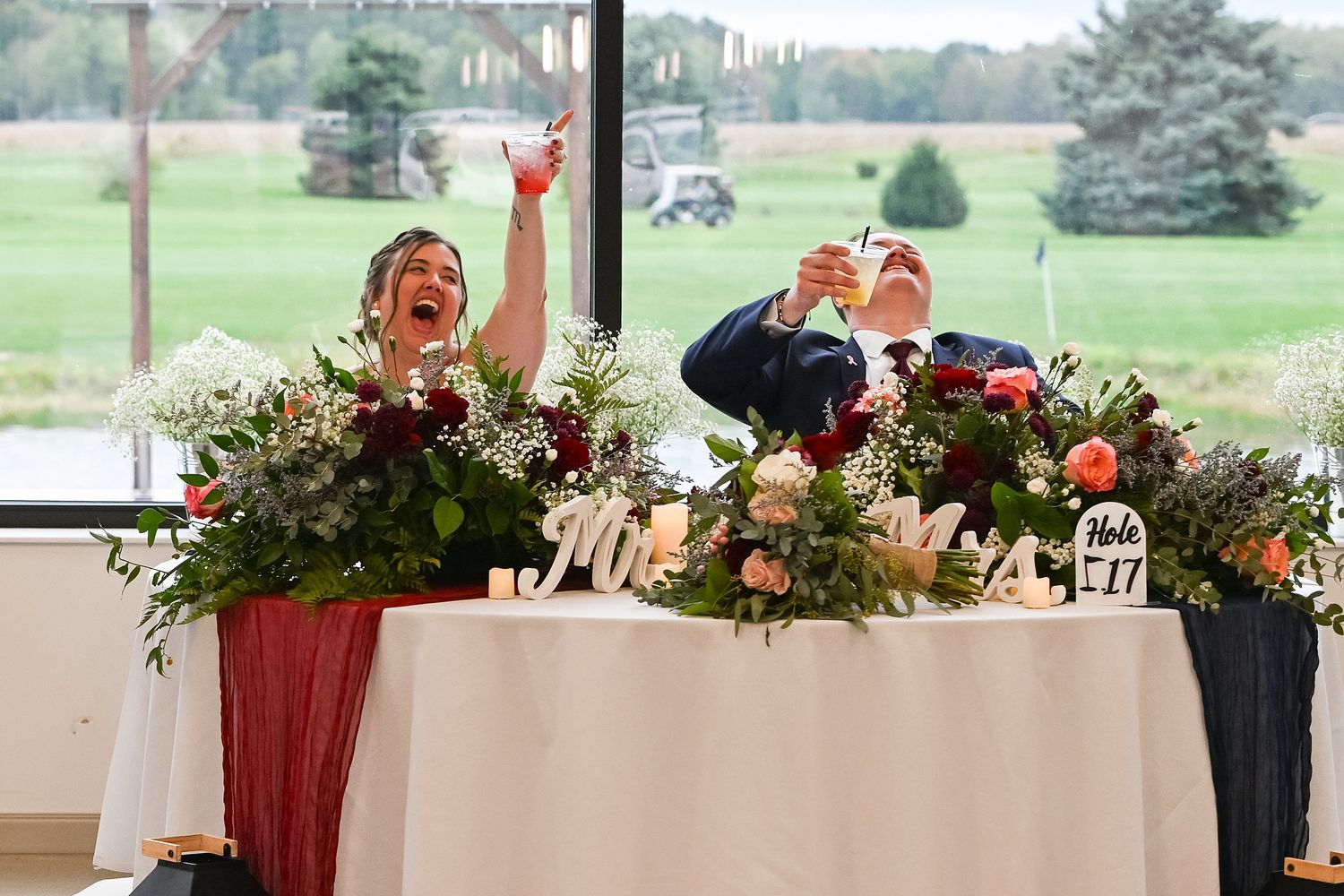 Bride and groom laughing joyfully at sweetheart table decorated with lush floral arrangements during wedding reception.