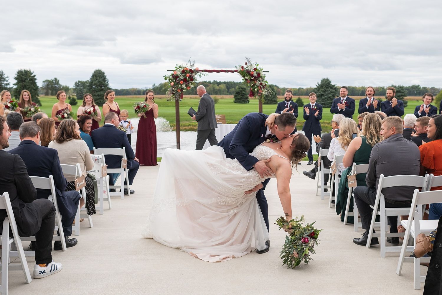 Groom dips bride for romantic kiss at outdoor wedding ceremony while guests applaud in white chairs.