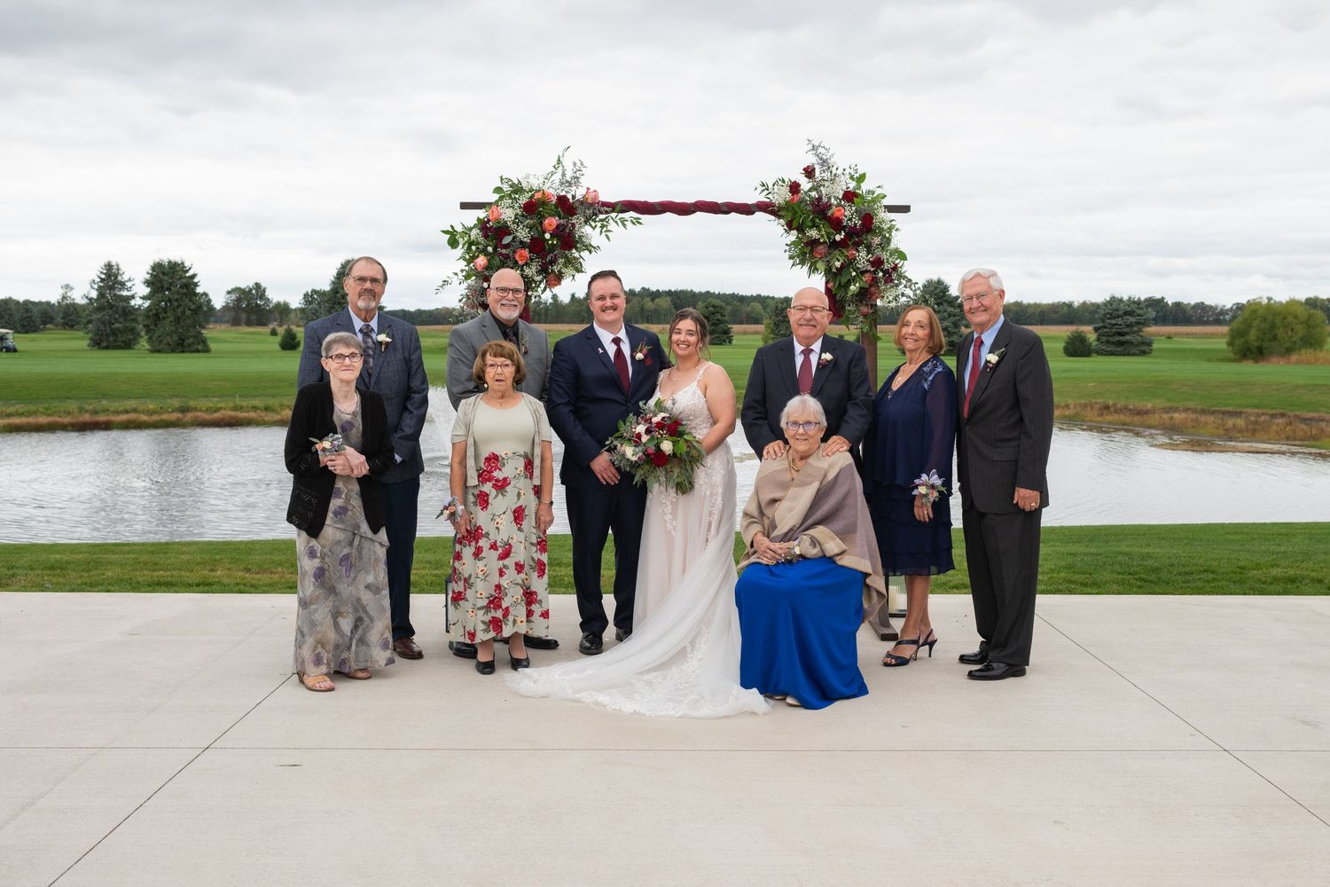 Wedding party poses under floral arch by pond, bride in white gown with groom and family members.