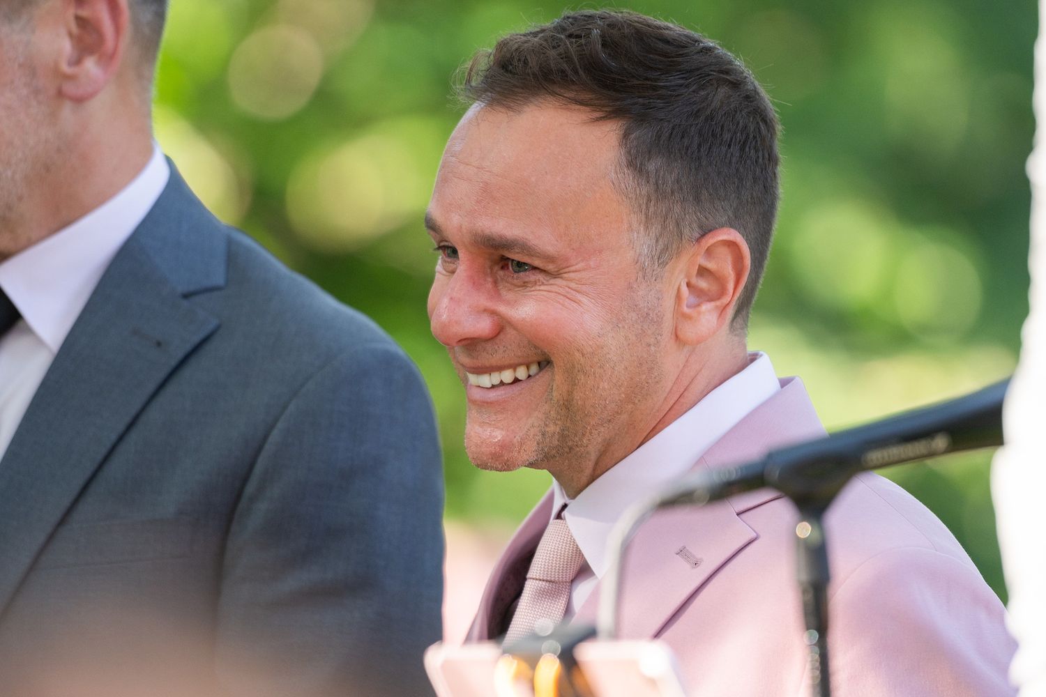 A person in a light pink suit jacket smiles warmly while standing outdoors with a blurred green background.