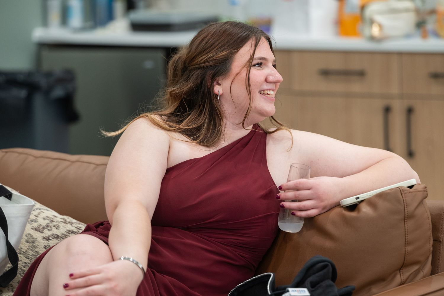 Woman in burgundy one-shoulder dress smiling while relaxing on a brown couch at a party.