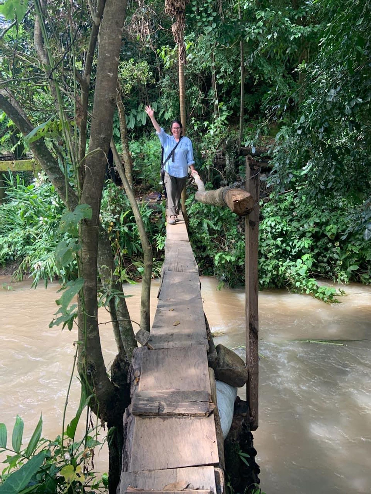 Person crossing a rustic wooden footbridge over a river surrounded by lush tropical vegetation.