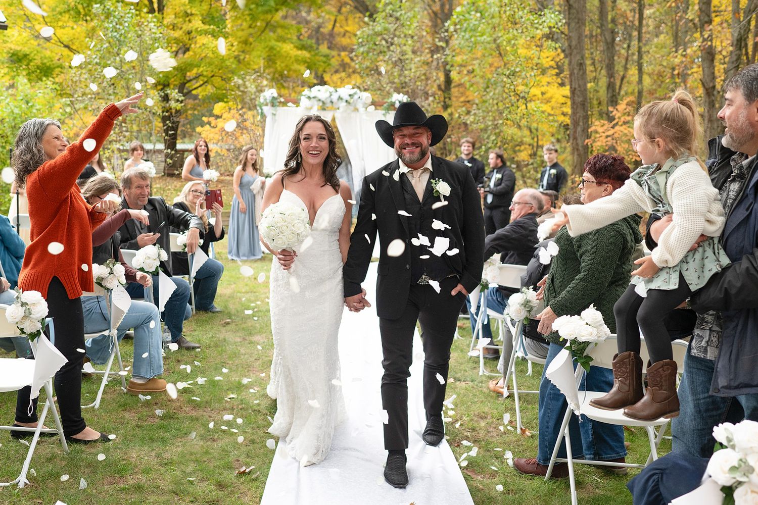 Newlyweds walking down outdoor aisle while guests throw confetti in autumn setting.