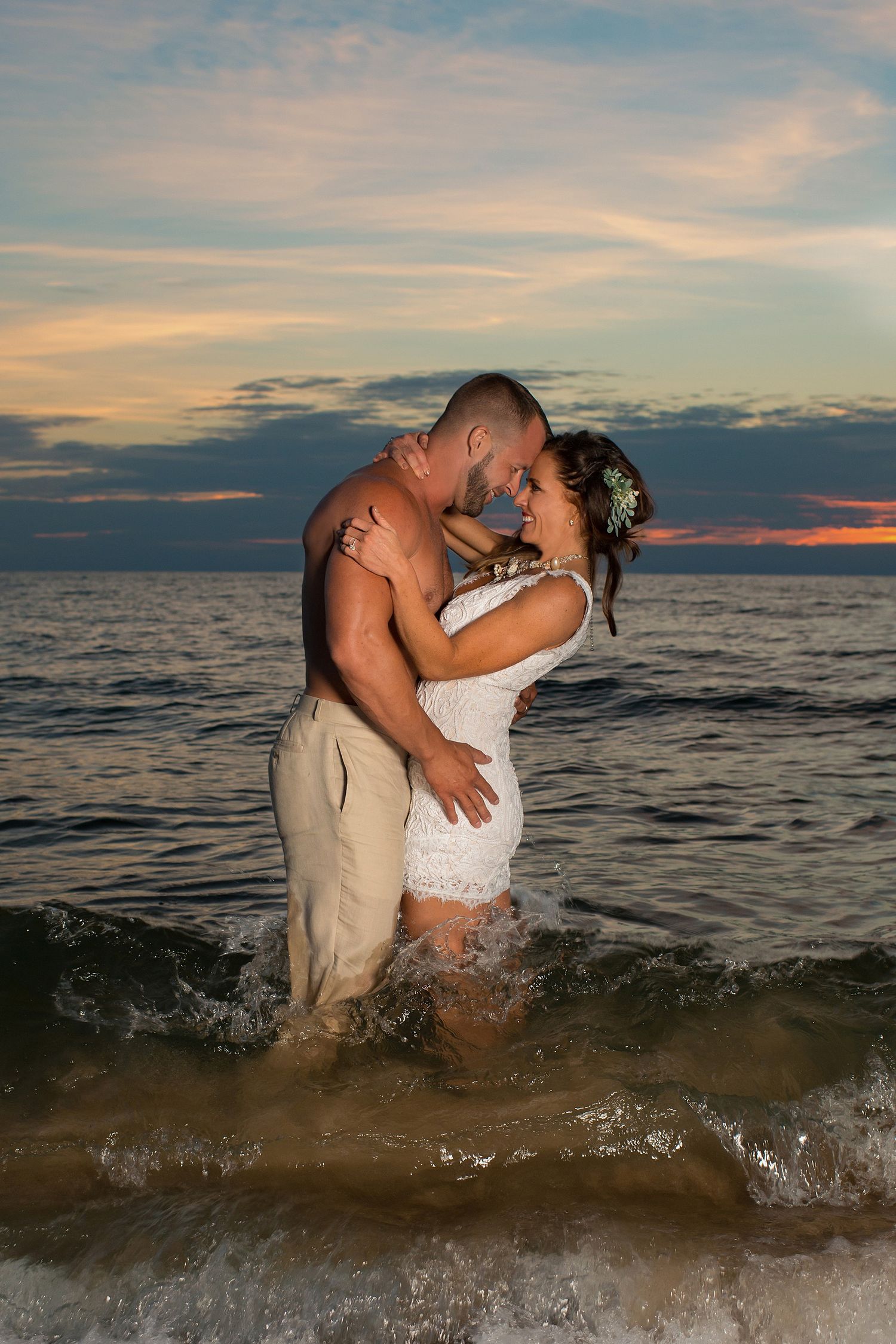 Couple sharing a romantic kiss in ocean waves at sunset, bride in white lace dress with floral hair accessory.