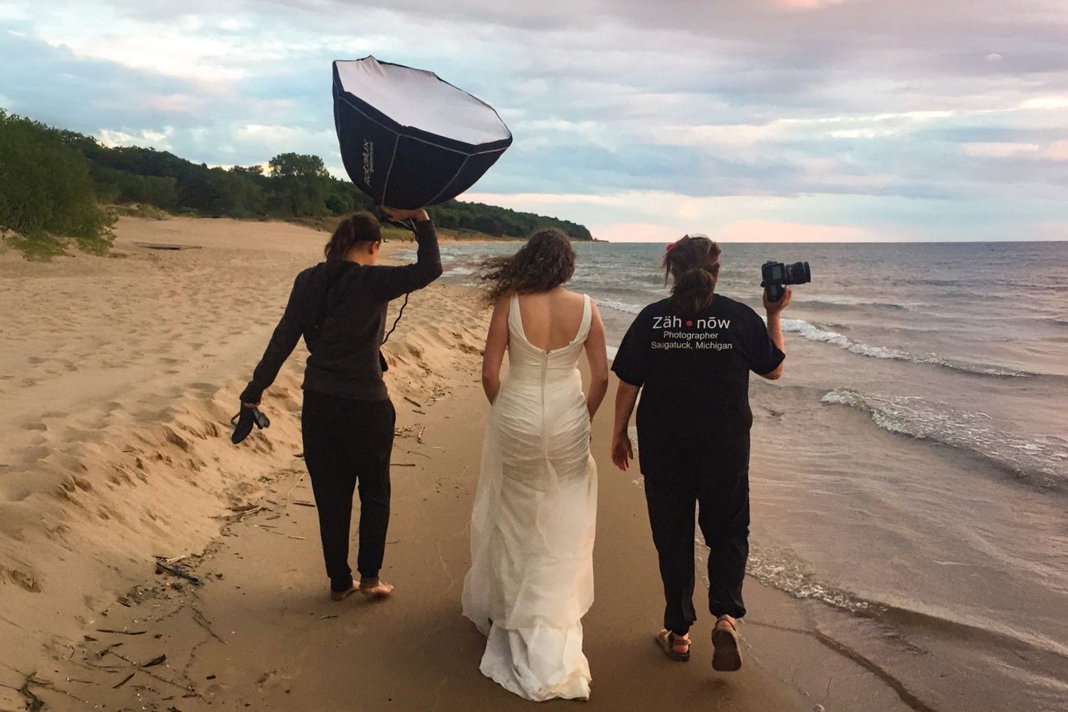 Three people walking along a beach at sunset with photography equipment and a white dress.