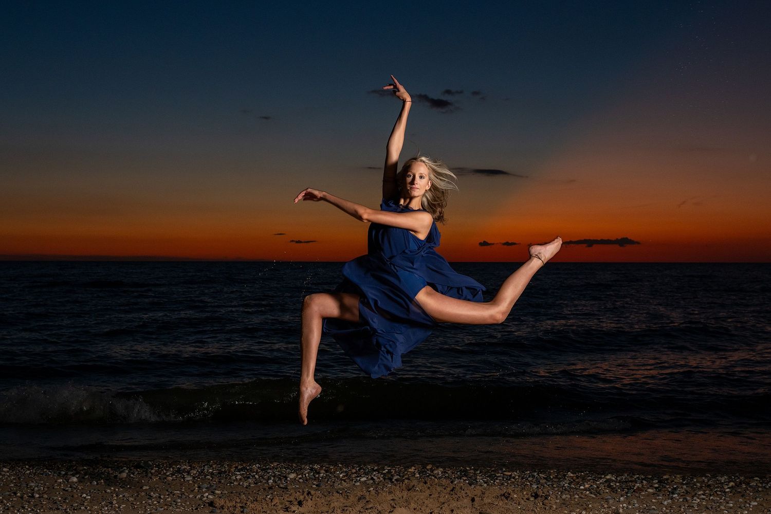 Athletic dance leap photographed against deep orange sunset sky over ocean waves.