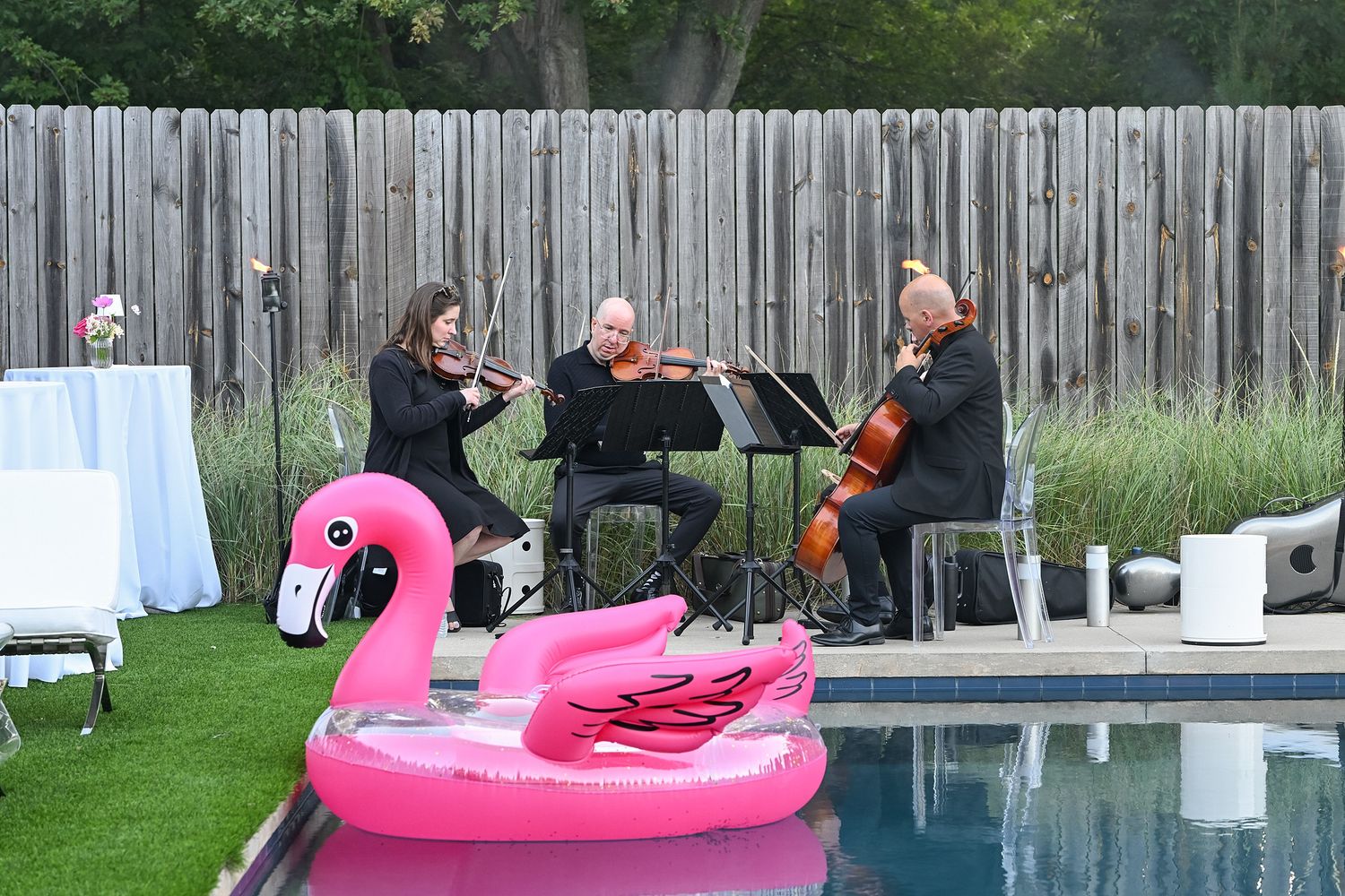 People enjoying a summer pool party with pink flamingo floats and wooden fence backdrop.