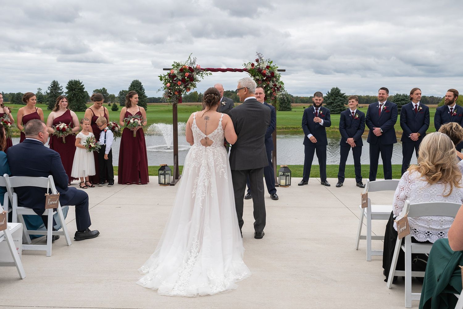 Bride and escort walk toward floral arch altar with wedding party lined up outdoors.