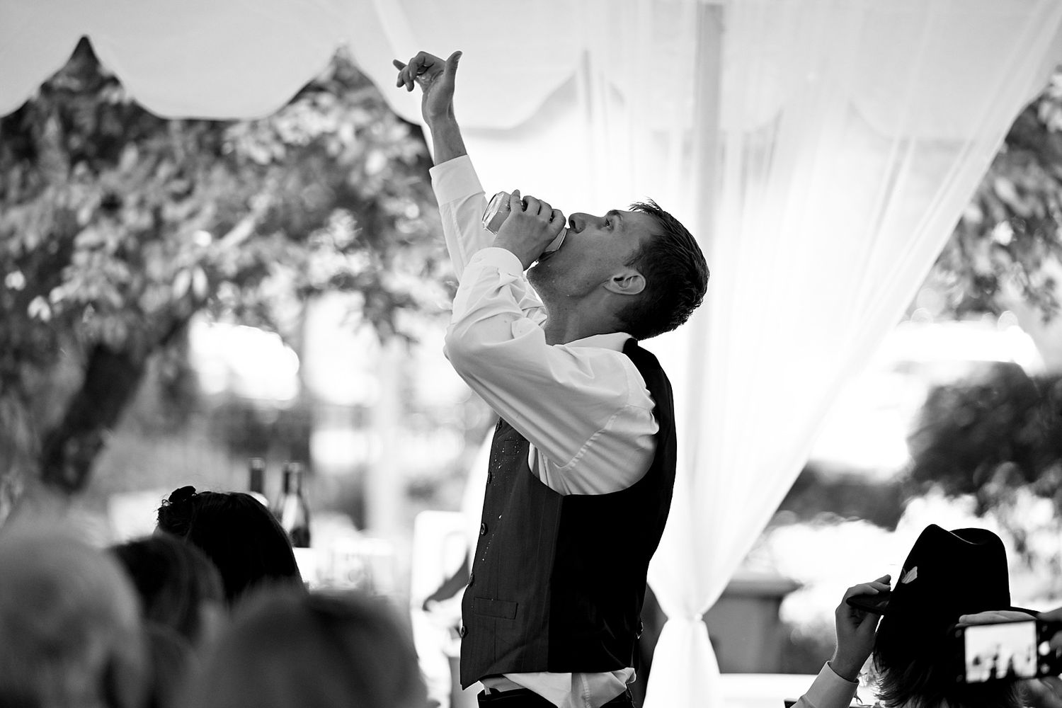 Black and white photo of a groom in a vest raising his arm at an outdoor wedding reception under a white tent.