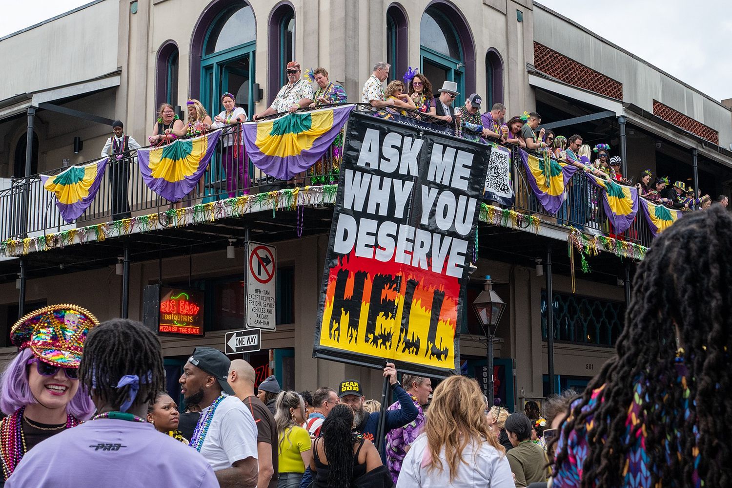Vibrant Mardi Gras decorations and signs hanging from balconies along a historic New Orleans street.