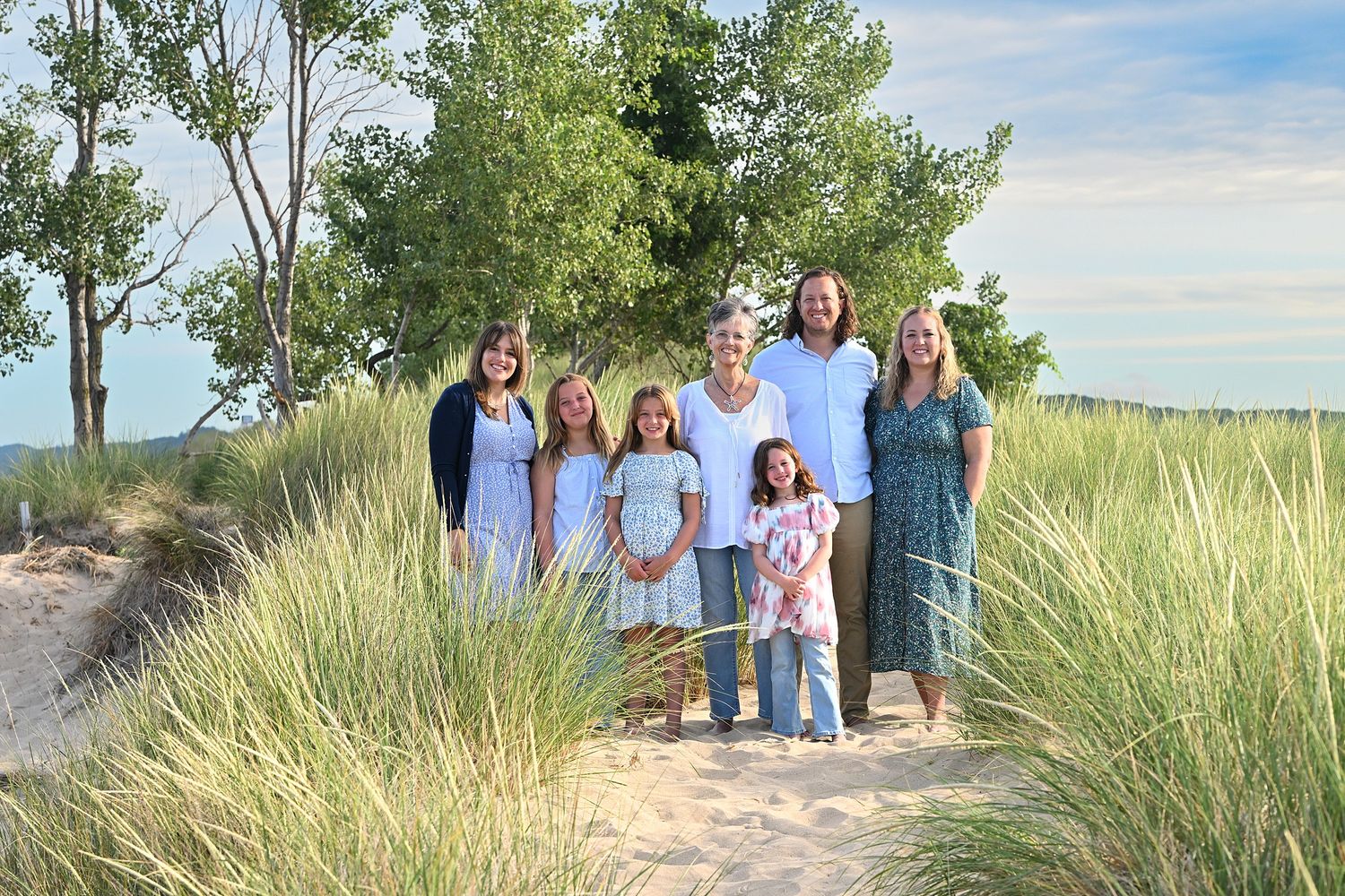 Family group of women and girls posing on sandy beach dunes with green grass and trees in background.