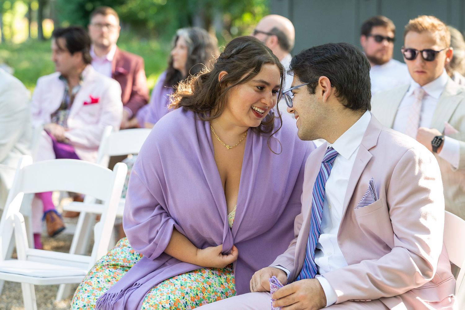 Couple in purple and white outfits share intimate moments at an outdoor event with guests seated in background.