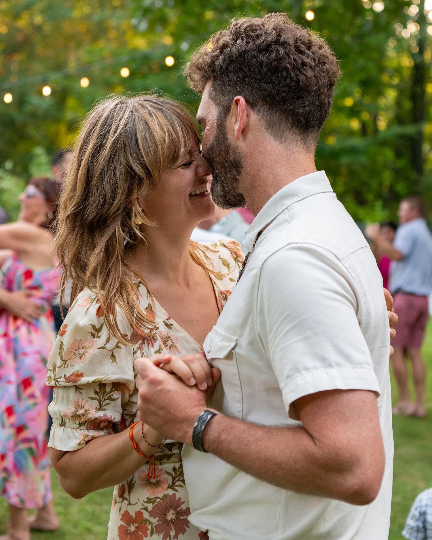 A couple dances together outdoors at a summer event with string lights and greenery in the background.