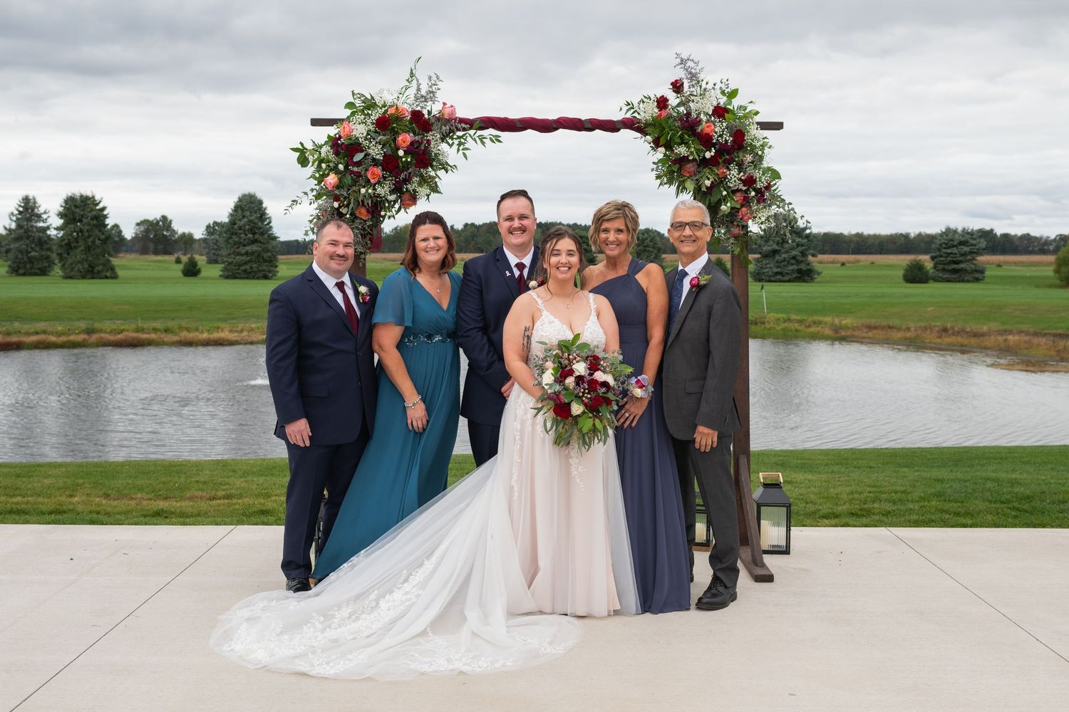 Wedding party poses under floral arch by pond, bride in white gown with bouquet, guests in formal attire.