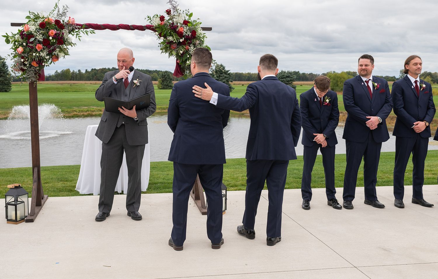 Outdoor wedding ceremony with groomsmen in navy suits standing near a floral arch by a pond.